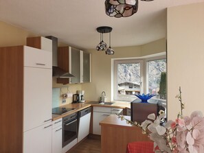 Una cuina moderna amb armaris blancs, un rentaplats, un forn, una cafetera i una finestra amb vista a cases i muntanyes.