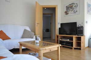 A living room with a white sofa, a wooden coffee table, a TV on a wooden stand, and a door leading to another room.