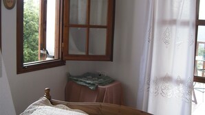 Un dormitorio con una cama de madera, una ventana con marco de madera, una mesita con un mantel verde y una cortina blanca con estampado floral.