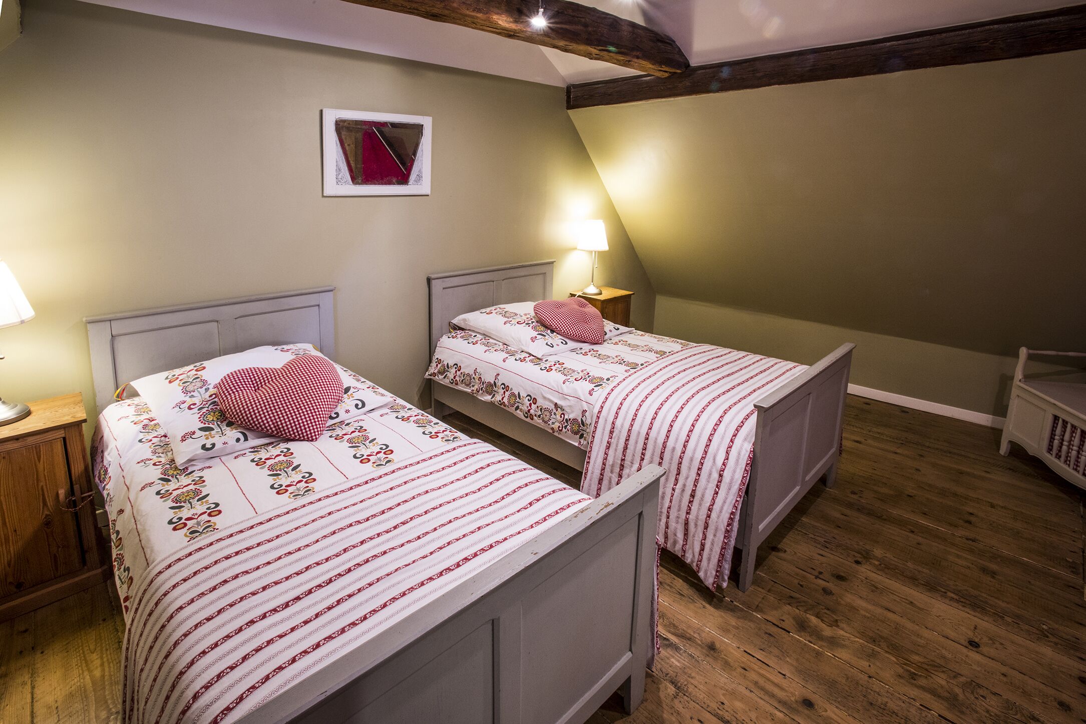 Une chambre dotée de deux lits simples, chacun avec une tête de lit, une table de chevet et une lampe. La chambre a un plancher en bois et un plafond en pente avec des poutres apparentes.