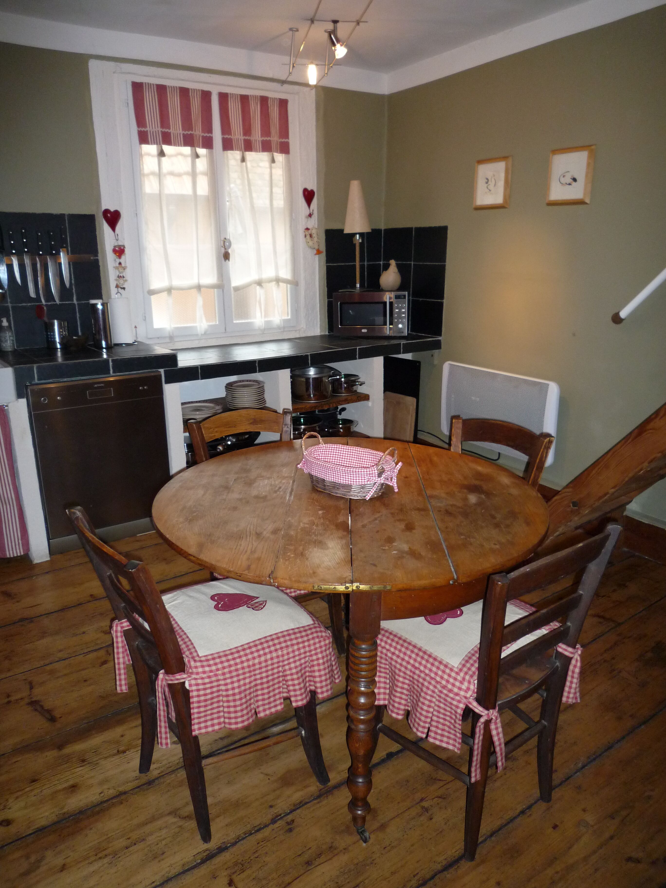A dining area with a round wooden table, four chairs, and a checkered tablecloth. There is a window with curtains and a microwave on the counter.