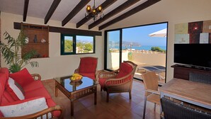 Een woonkamer met een rode bank, twee rieten fauteuils, een glazen salontafel, een televisie en uitzicht op de zee.