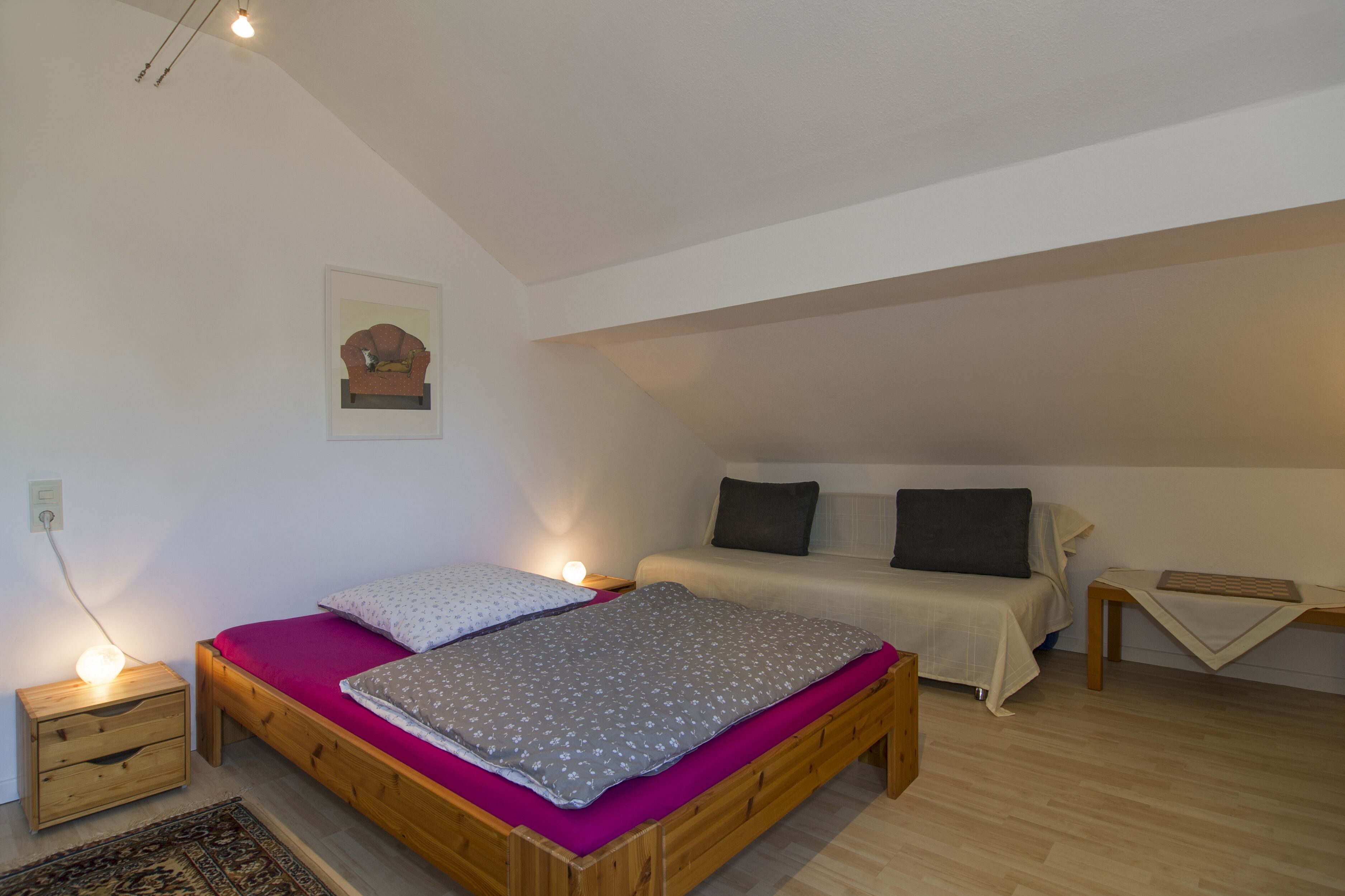 A bedroom with a wooden bed, a nightstand with a lamp, a small table with a keyboard, and a wall-mounted picture.