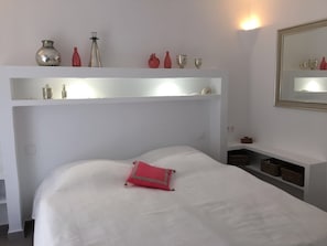 Una cama bien hecha con una colcha blanca y una almohada roja, una pared blanca con una repisa que sostiene varios objetos decorativos, y un espejo en la pared a la derecha.