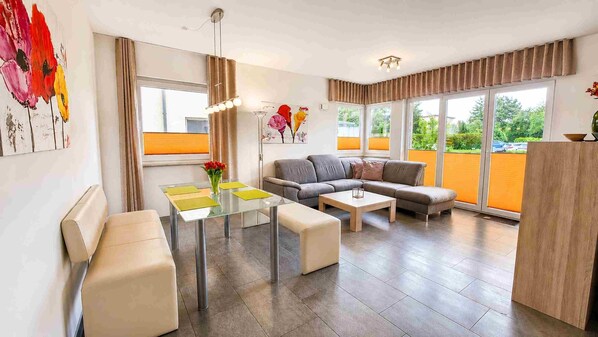 A modern living room with a sofa, a glass dining table, and a view of greenery outside. - Apartment AS04 - Villa hill (Heringsdorf)