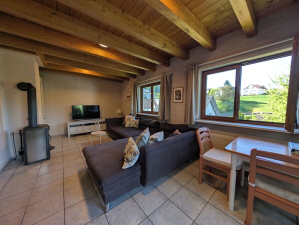 A cozy living room with a dark sofa, a wooden table, a TV, and a large window with a view of greenery and a house.