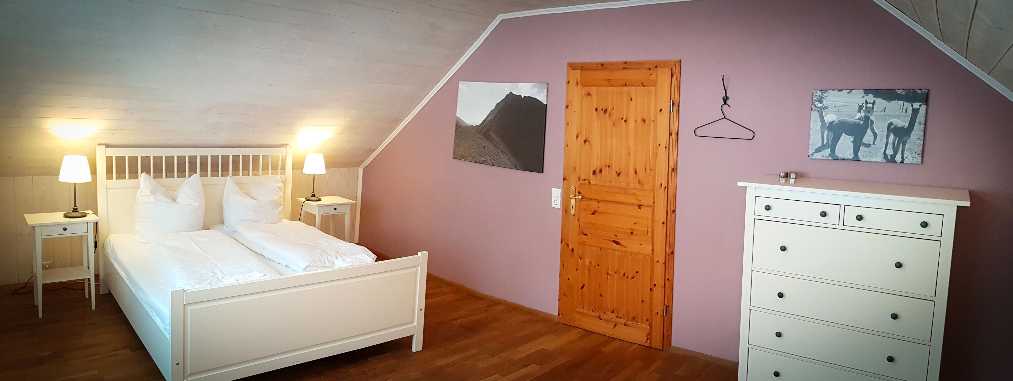 Une chambre à coucher comprenant un lit, des tables de chevet, une commode, une porte en bois et deux tableaux accrochés au mur.