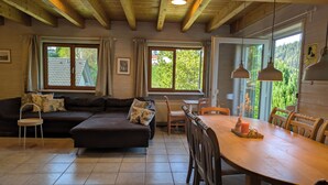 Een gezellige woonkamer met een hoekbank, een eettafel en stoelen. Grote ramen zorgen voor natuurlijk licht en uitzicht op groen buiten.