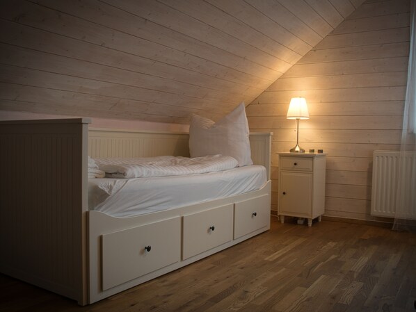 Une chambre avec un plafond en pente, un lit avec une literie blanche, une table de chevet avec une lampe et un plancher en bois.