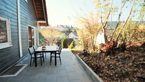 Een terras met een tafel en stoelen, een klein tuinperk met planten en een boom, en een huis op de achtergrond.