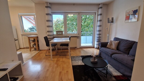 Una sala de estar moderna con un sofá, una mesa de comedor con sillas, una pequeña mesa de centro y vistas amplias a través de grandes ventanales.
