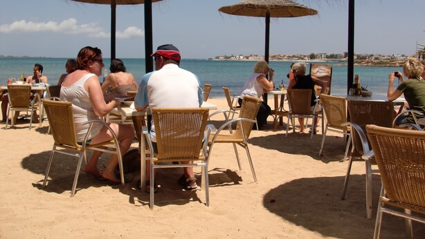 Un restaurant situé en bord de mer, avec une terrasse comprenant des chaises et des tables en osier, ainsi que des parasols en chaume pour offrir de l’ombre.