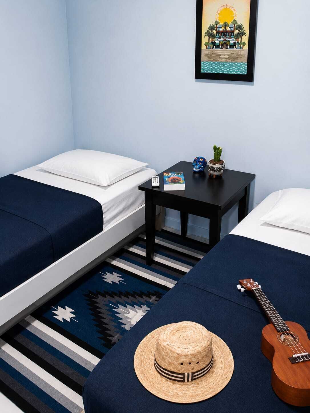 A room with two beds, a small table with a book and a plant, a guitar, and a framed picture on the wall.