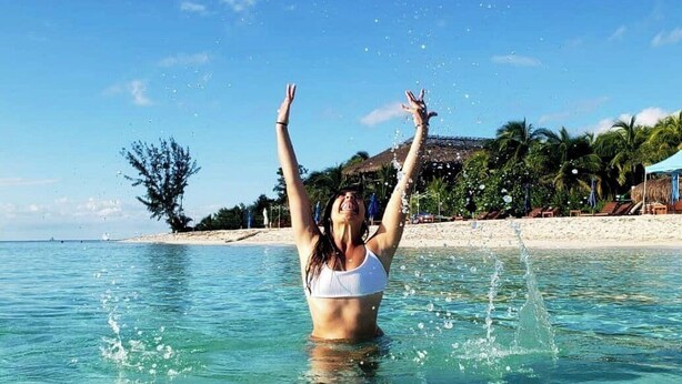 Una persona en traje de baño en agua cristalina, con una playa y palmeras al fondo.