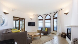 Un salon spacieux comprenant un canapé, des fauteuils, une table basse et une cheminée. De grandes fenêtres, ornées de rideaux, laissent entrer la lumière, et un téléviseur à écran plat est fixé au mur. La vue donne sur l’extérieur.