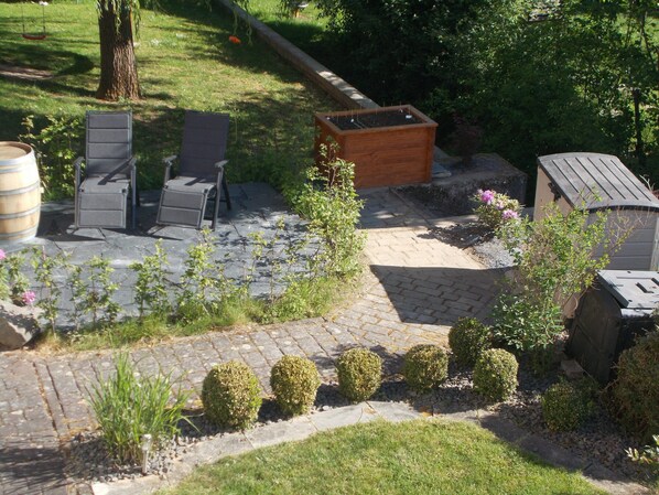 Un espace patio doté d’un sol en pierre, entouré de petits arbustes et agrémenté d’un bac à fleurs en bois.