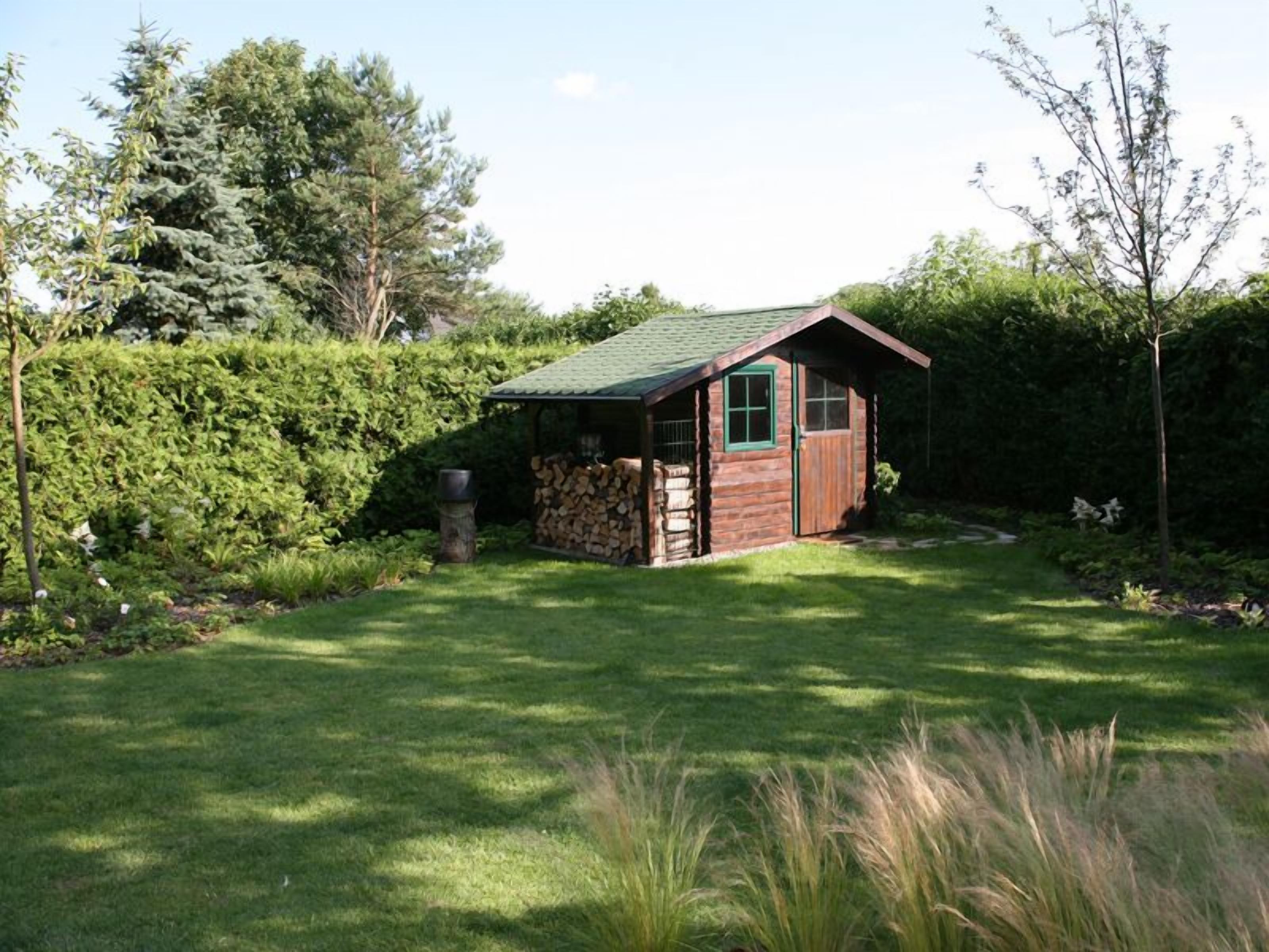 Una pequeña cabaña de madera con techo verde, rodeada de un césped cuidado y abundante arbustal.