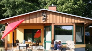 Et træhus med en overdækket terrasse, et bord, stole og en rød parasol.
