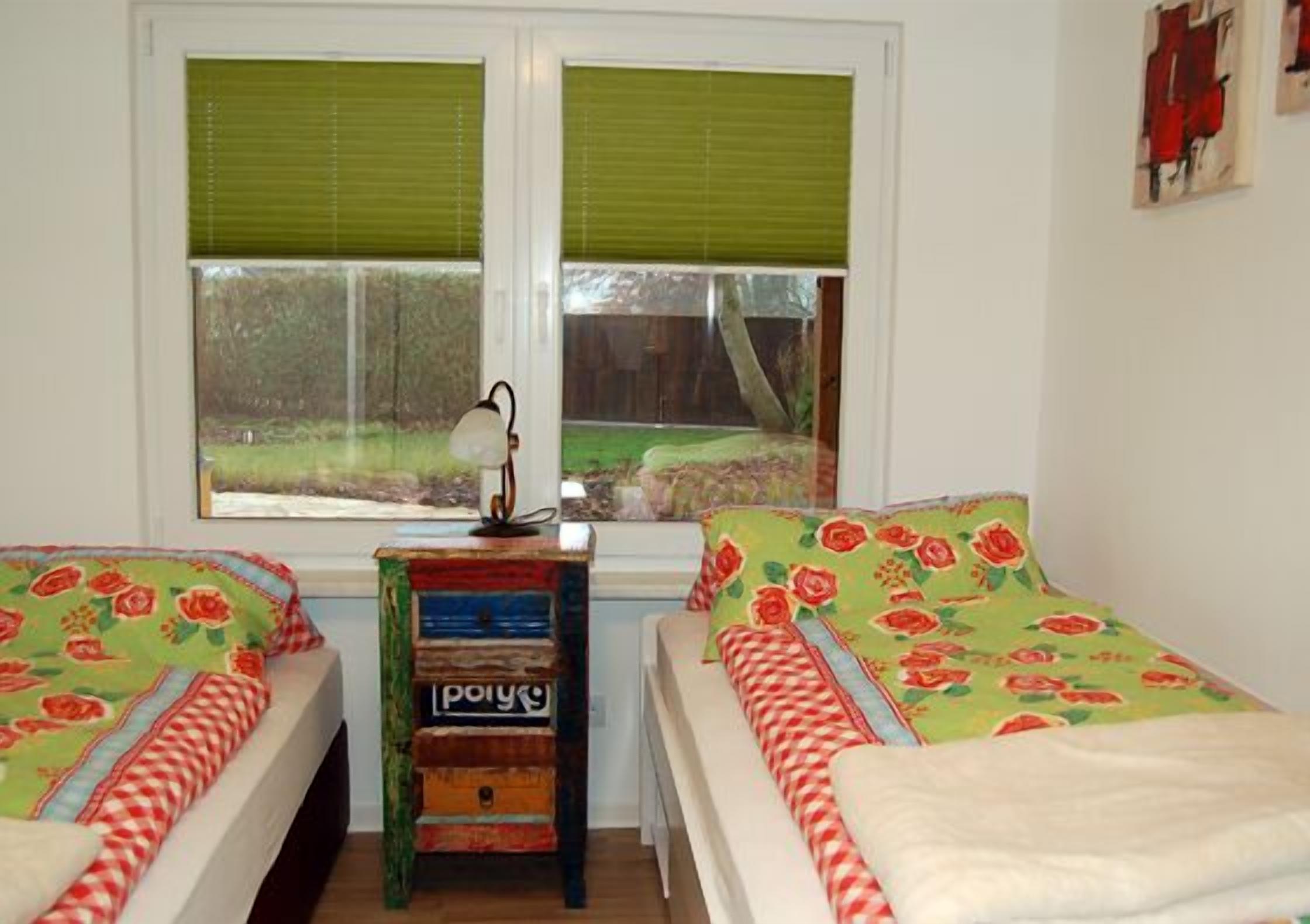 A room with two beds, a small table with a lamp, and a window with blinds.
