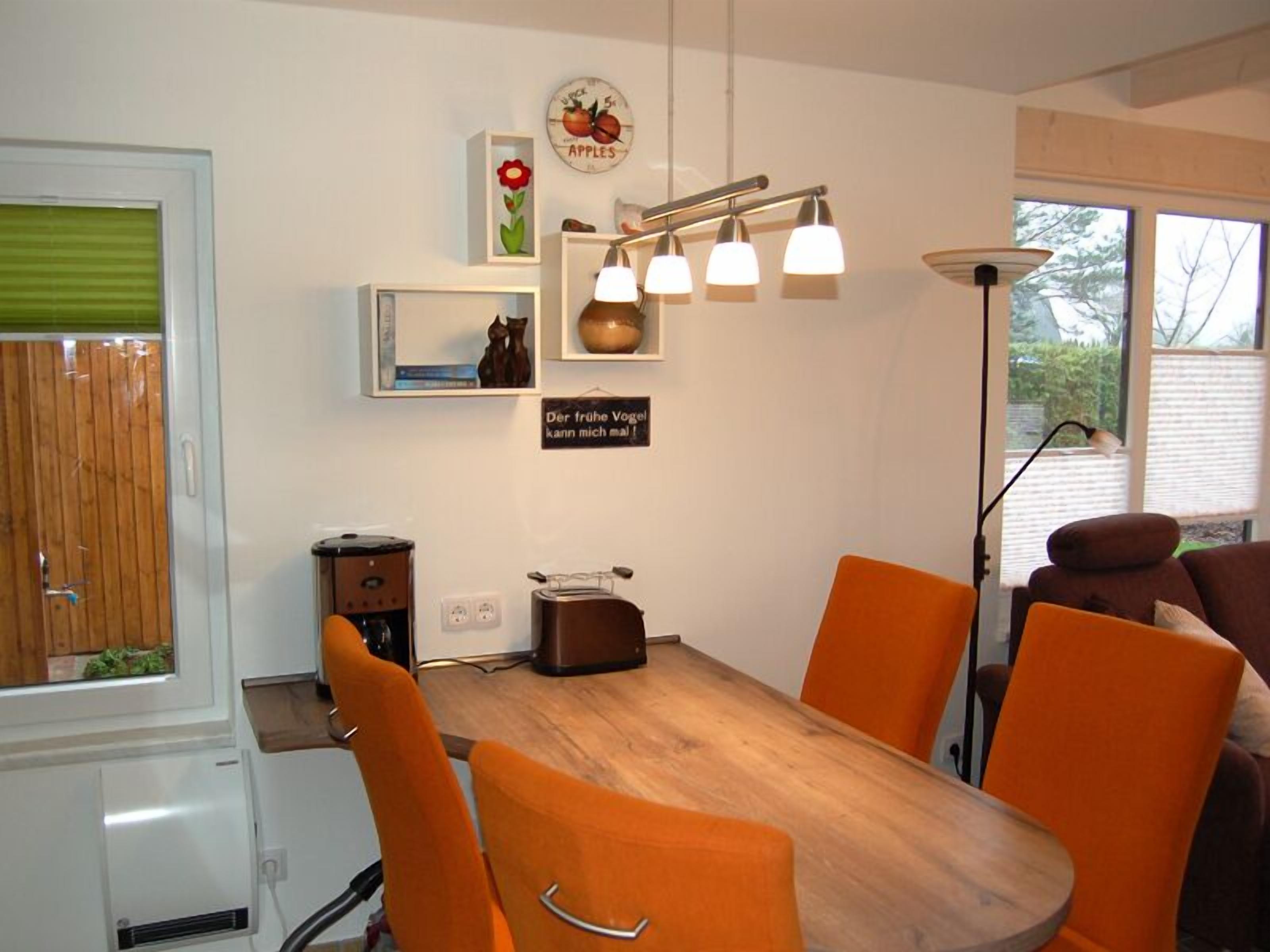 A dining area with a wooden table and orange chairs, a coffee machine, and a wall-mounted light fixture.