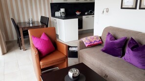 A compact living space with a brown leather chair, a sofa with purple cushions, a small coffee table, and a kitchenette in the background.