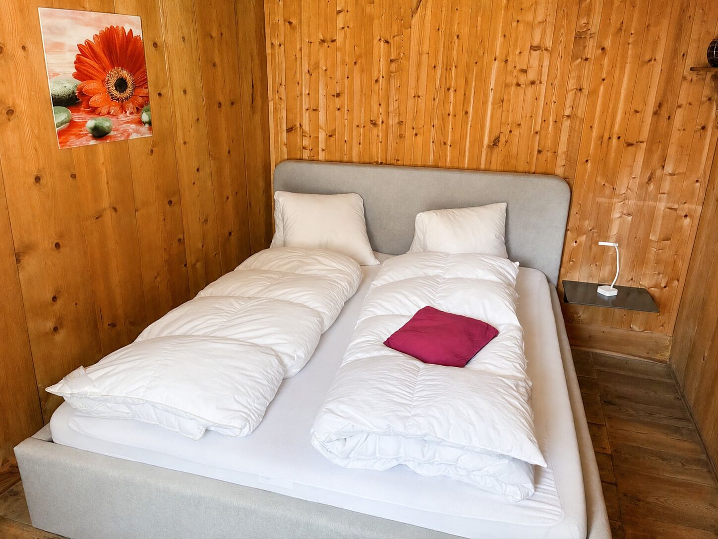 A single bed with white bedding and a red pillow, set against a wooden wall with a framed picture.
