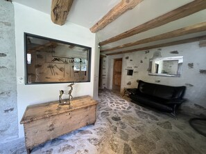 A rustic room with a stone floor, wooden beams, a wooden bench, a black leather sofa, and a large mirror.