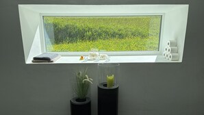 Une fenêtre donnant sur un champ, deux vases avec des plantes et un petit rebord de fenêtre sur lequel se trouvent une serviette et un canard en caoutchouc.