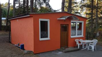 A small orange building with a single door and window, a small porch with a table and chairs, and a blue object on the ground.
