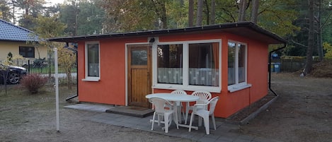 Un pequeño edificio de una sola planta con fachada roja y detalles blancos, que presenta una puerta de madera y ventanas amplias. Cuenta con un pequeño patio con una mesa y sillas.