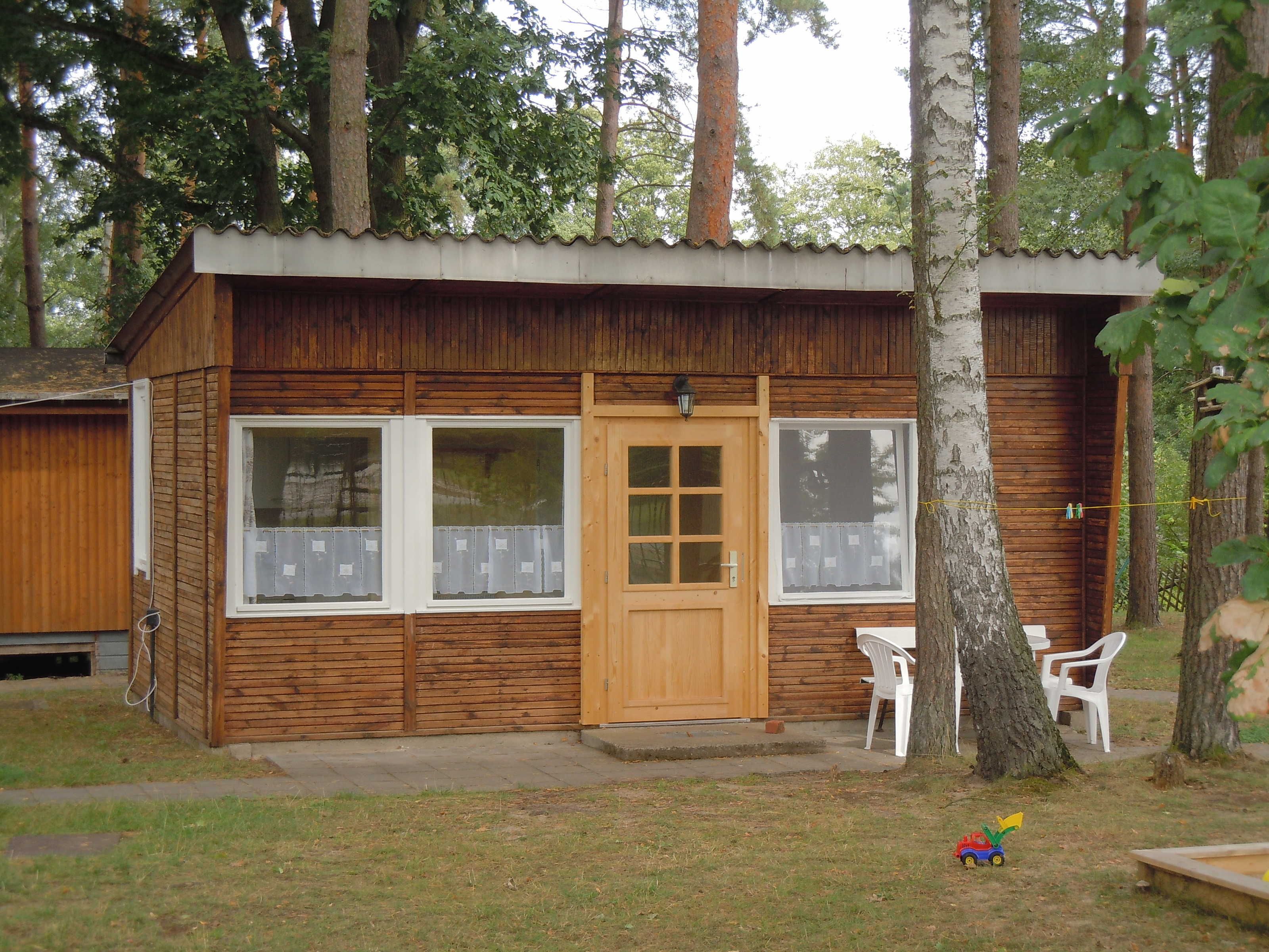 Una cabana de fusta amb una porta d'entrada, dues finestres i un petit porxo amb cadires de plàstic.
