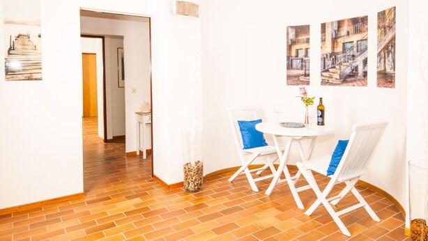 Una habitación luminosa con paredes blancas, piso de ladrillo y un pequeño comedor con sillas y mesa blancos.