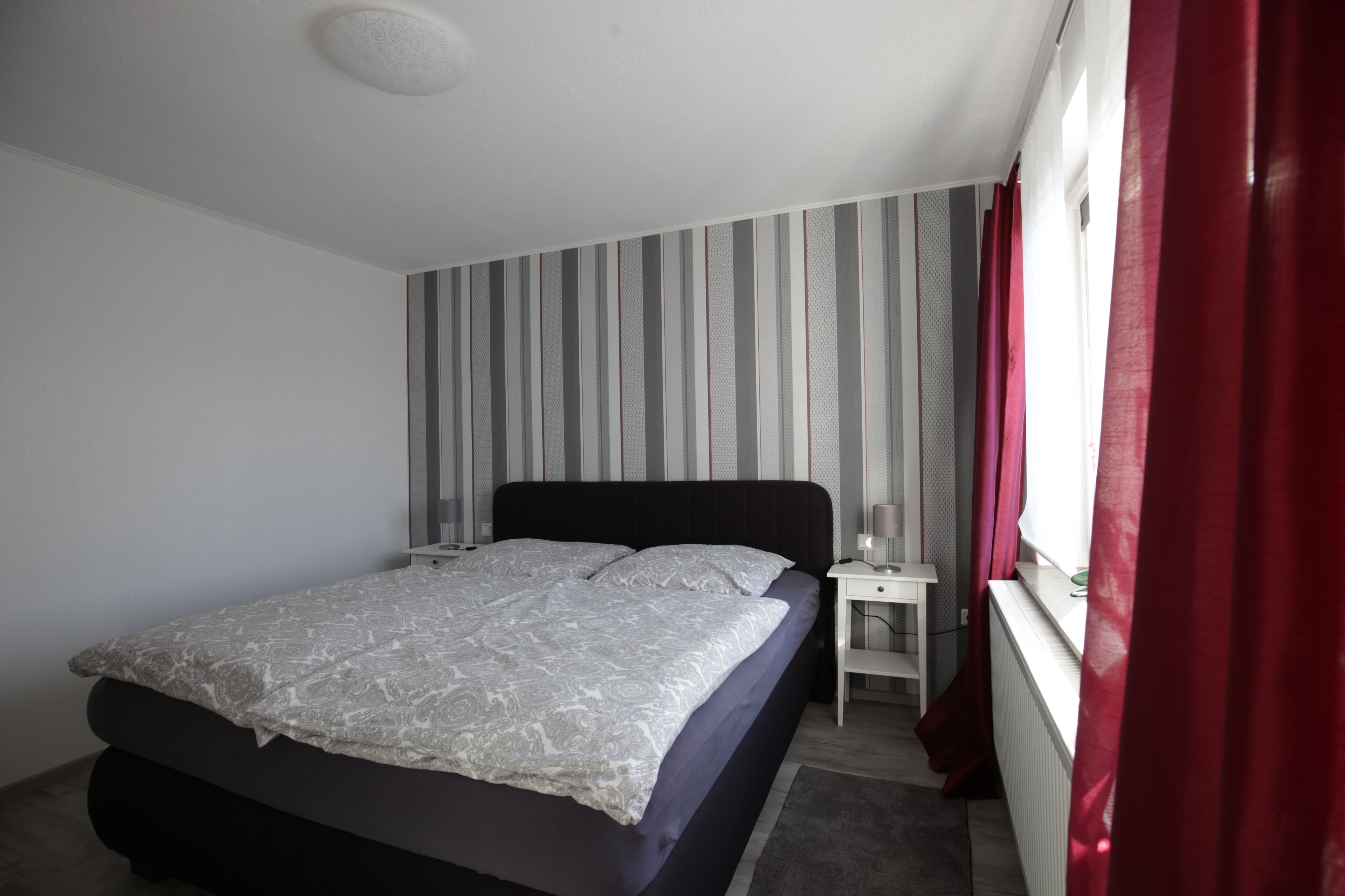 Quarto com cama, mesa de cabeceira, janela com cortinas e parede listrada.