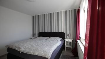 Quarto com cama, mesa de cabeceira, janela com cortinas e parede listrada.