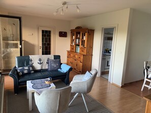 Un salon avec un canapé bleu foncé, un fauteuil gris, un meuble en bois et une cuisine visible à travers une porte ouverte.