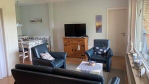 Una sala de estar con un sofá, dos sillones, un televisor sobre un mueble de madera, una zona de comedor con una mesa y sillas, y una ventana con vistas.