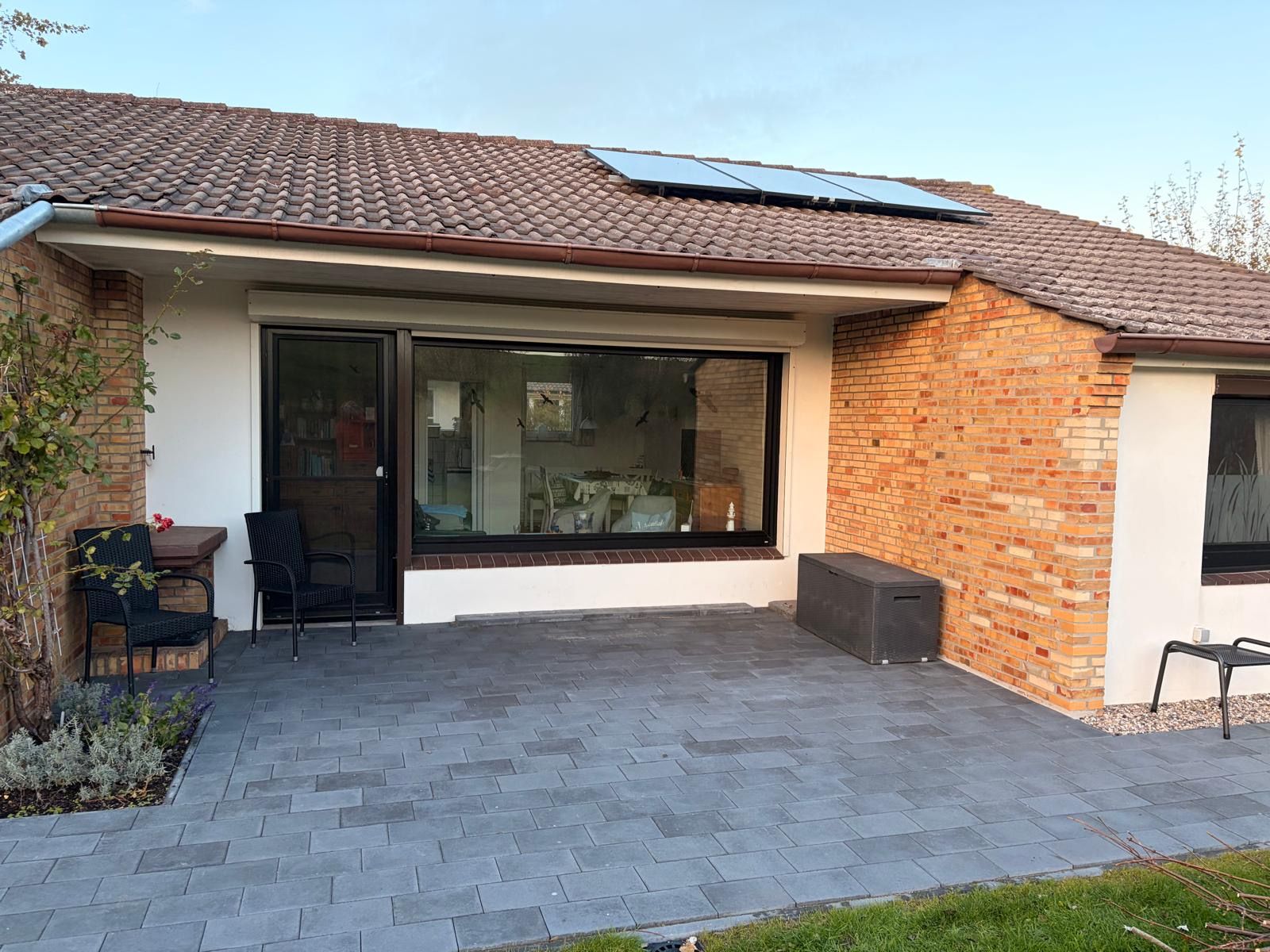 A residential building with a tiled roof, a large sliding glass door, a brick exterior, and a paved patio area with outdoor furniture.