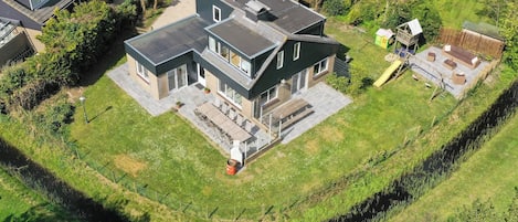 A modern two-story house with a large patio area, a small playhouse, and a fenced yard.