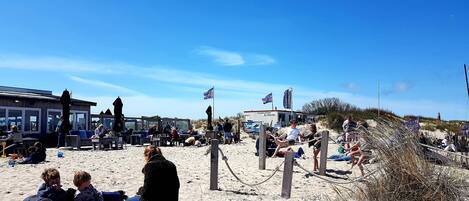 Una playa con gente relajándose, un edificio con banderas y un faro al fondo.