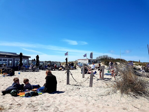 A beach with people relaxing, a building with flags, and a lighthouse in the background.