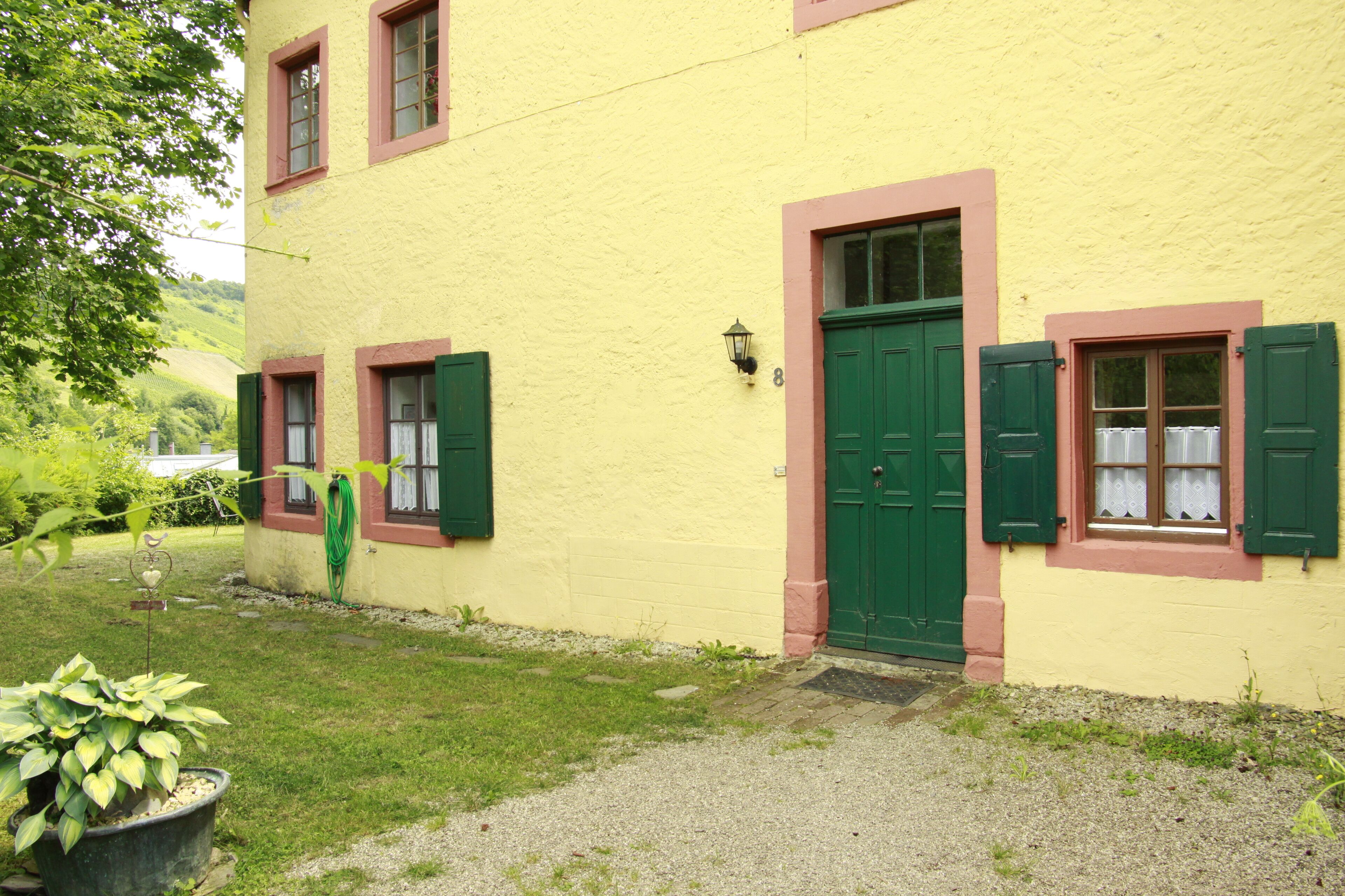 Un bâtiment jaune avec des volets verts et une porte verte, au numéro 8, avec un jardin devant.