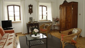 A living room with a sofa, a television, a table with a vase, wicker chairs, a wooden cabinet, and two windows with curtains.