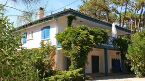 A two-story house with a blue roof and white walls, surrounded by greenery and trees. - Pleasant family villa, in the heart of Cap Ferret, close to the beach, 12 pers (Lège-Cap-Ferret)