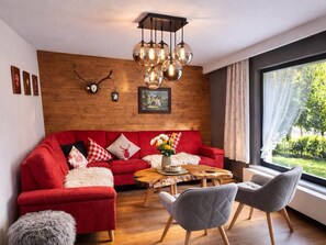 A modern living room with a red sofa, grey chairs, a wooden coffee table, and a large window with a view of greenery. - Alpenperle vacation home at the foot of the Zugspitze (Grainau)