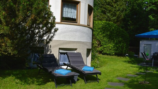Un bâtiment à l’extérieur blanc, avec une fenêtre ornée d’un rideau blanc. On y voit deux fauteuils noirs, sur lesquels sont posées des serviettes bleues.