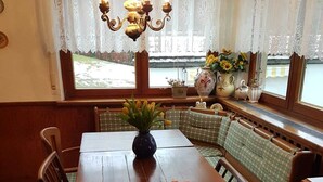 Un coin repas avec une table en bois, des bancs, un lustre et une fenêtre ornée de rideaux en dentelle.