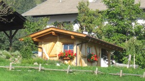 A wooden house with a sloped roof, a wooden porch, and flower boxes with red flowers. - Farm holidays Rotsteinhof (Vöran)