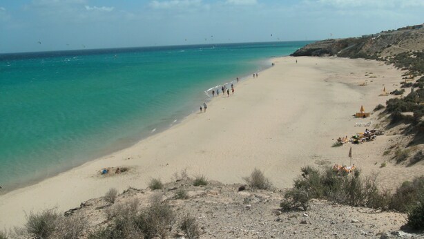 En strand med klart turkis vand, sandet bredde og sparsom vegetation.