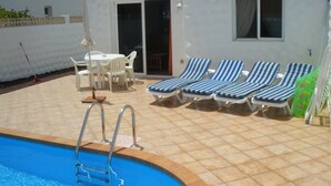En swimmingpool med stige, liggestole, et bord med stole, og en bygning med tagsten og vinduer.