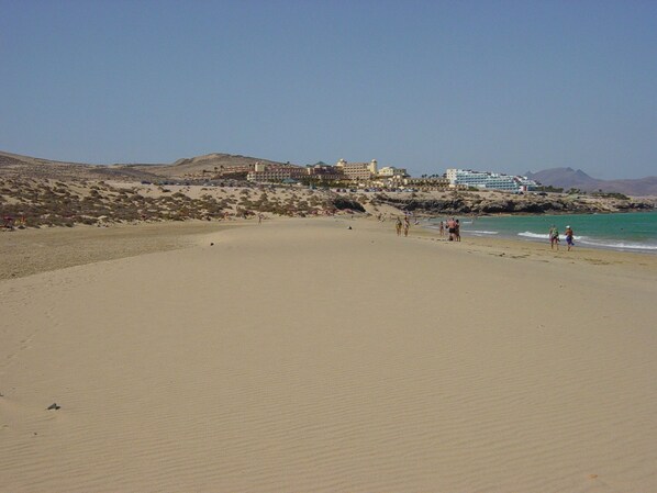 Ein weiter Sandstrand mit wenigen Spaziergängern, einer felsigen Küstenlinie und Gebäuden im Hintergrund.